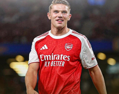 На изображении футболист в красно-белой форме футбольного клуба Арсенал. На переднем плане эмблема клуба и логотип спонсора Emirates Fly Better. Фон размытый, создающий атмосферу футбольного стадиона